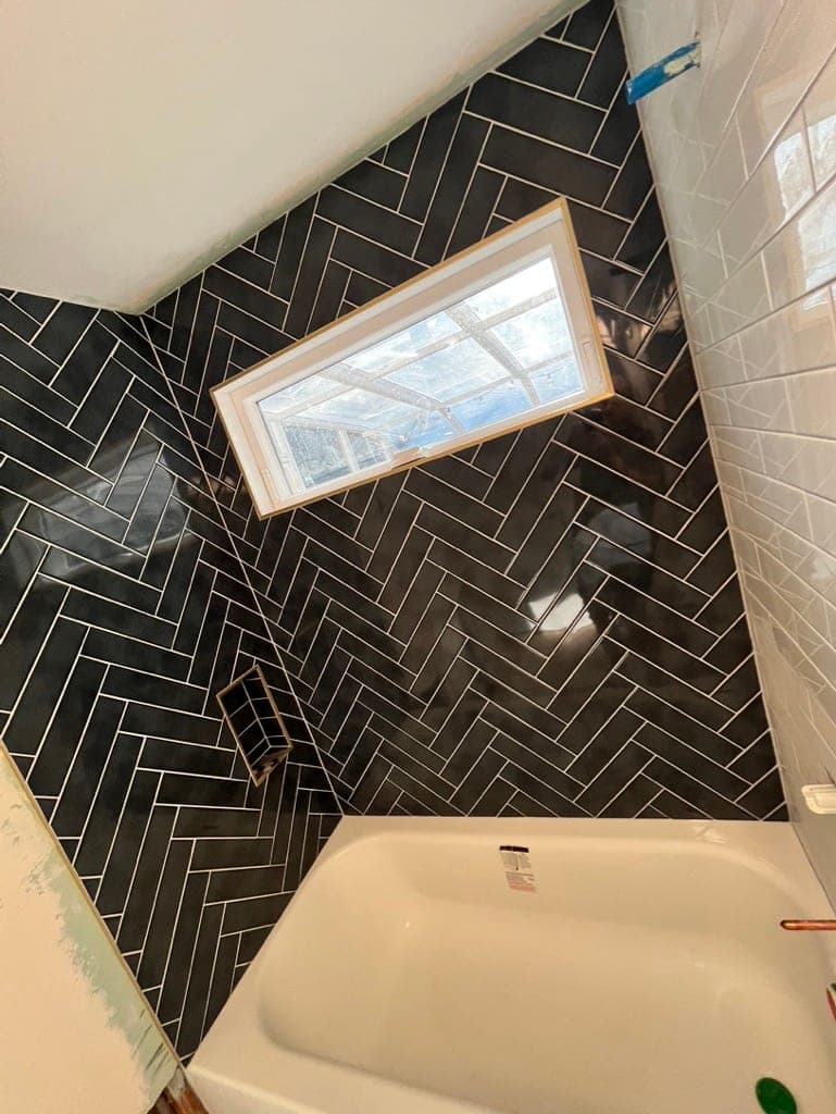Tub surround in glossy charcoal herringbone tile wrapped around a window, before grout