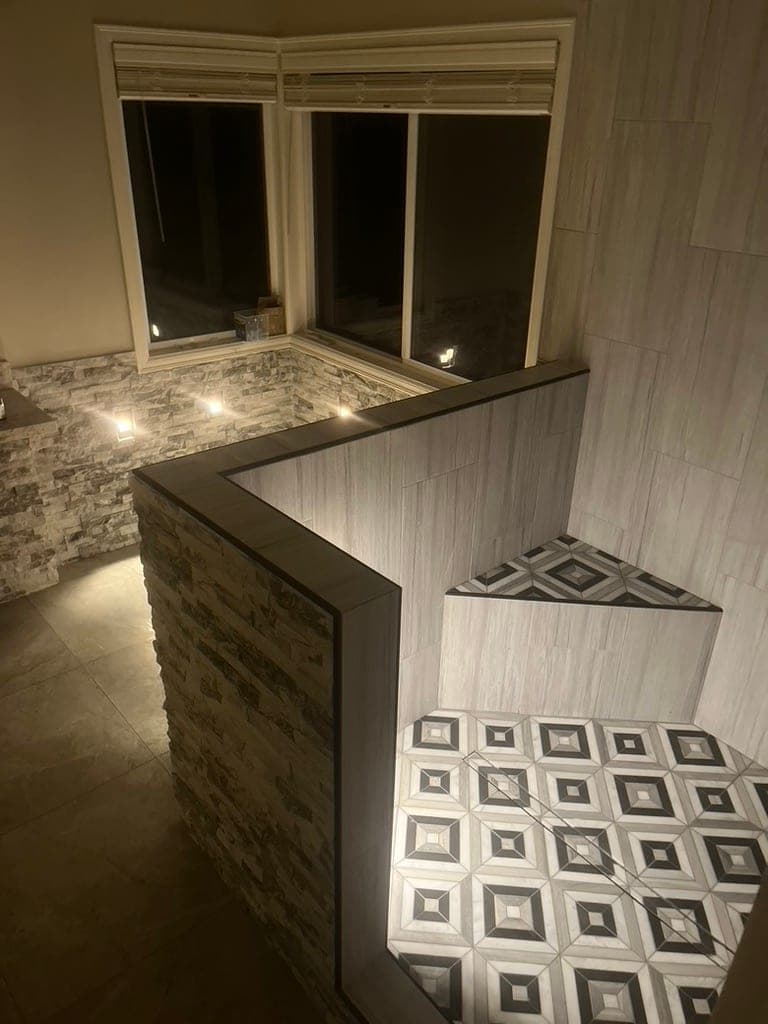Shower with gray wood-look walls, geometric mosaic floor, stacked-stone half wall, and lit niches