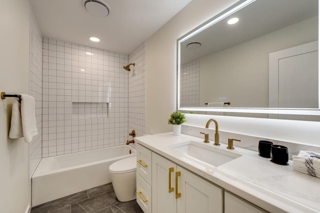 Bathroom with a white stacked tile tub surround, brass fixtures, backlit mirror, and gray plank floor